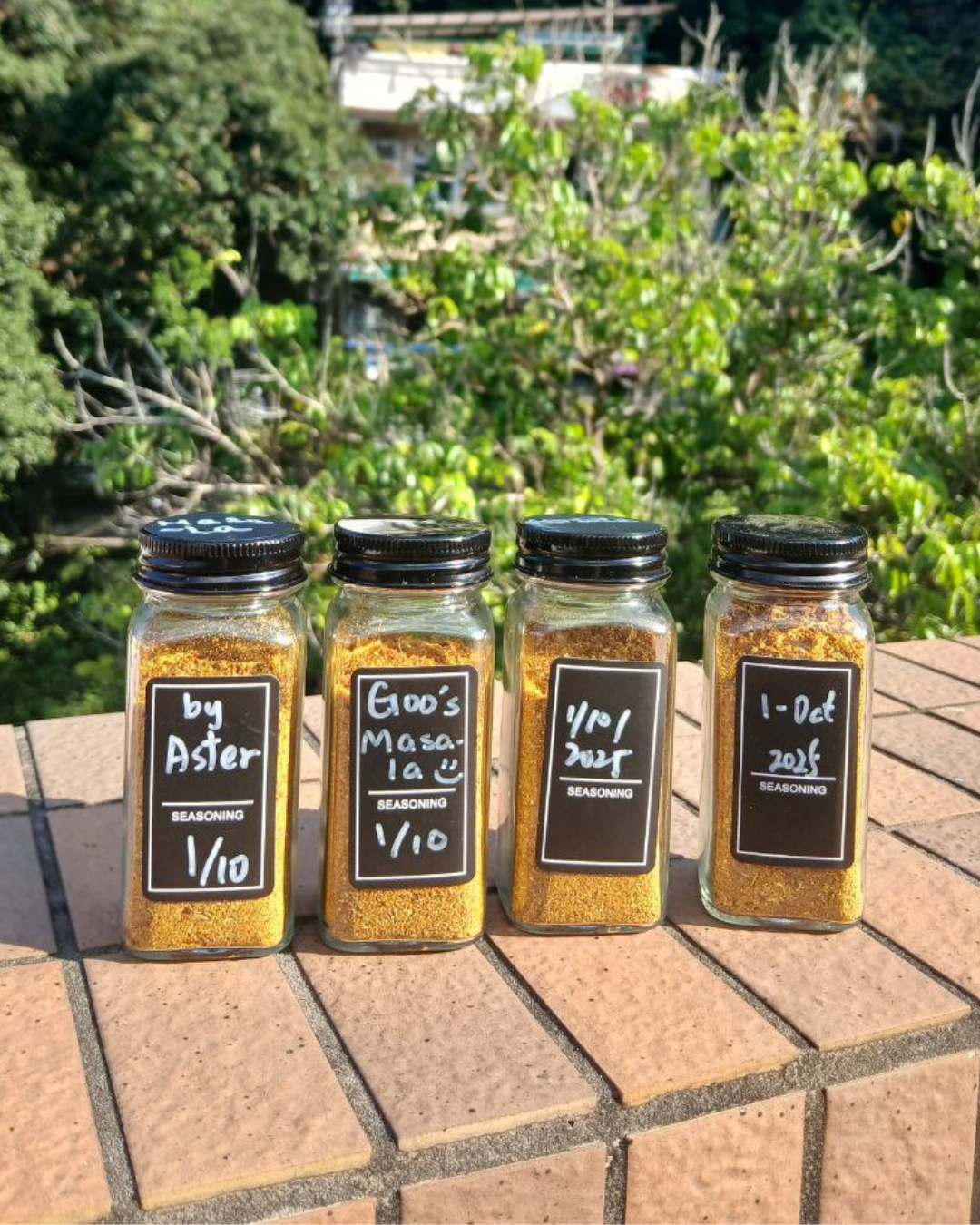 Four jars of seasoning with labels on a brick surface outdoors