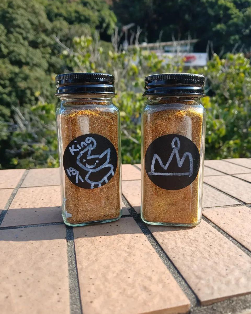Two glass jars of seasoning with black lids on a stone surface with a green outdoor background